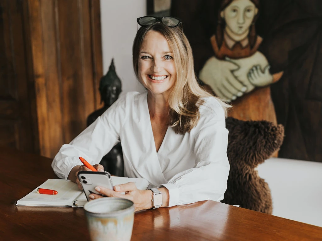 Heike Ott sitzt mit Handy und Notizblock an einem hölzernen Schreibtisch, im Hintergrund ein großes Gemälde